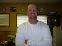 Smiling bald man in a white shirt standing indoors with arms crossed in a room with windows.