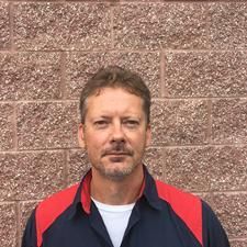 Man standing against a brick wall, wearing a navy jacket with red shoulders and a white shirt.