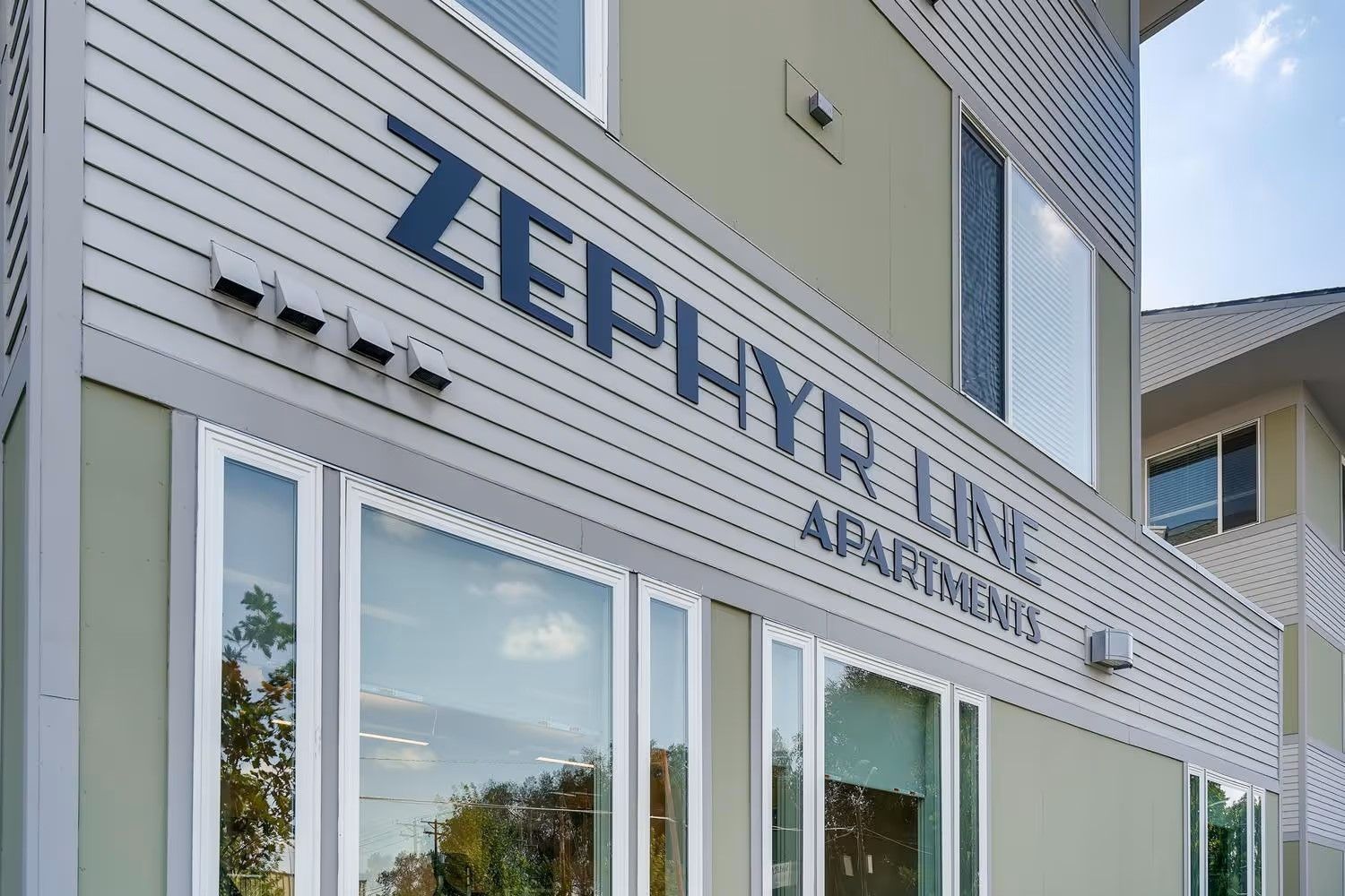 The exterior of the Zephyr Line Apartments building, featuring beige siding and large windows on a bright day.