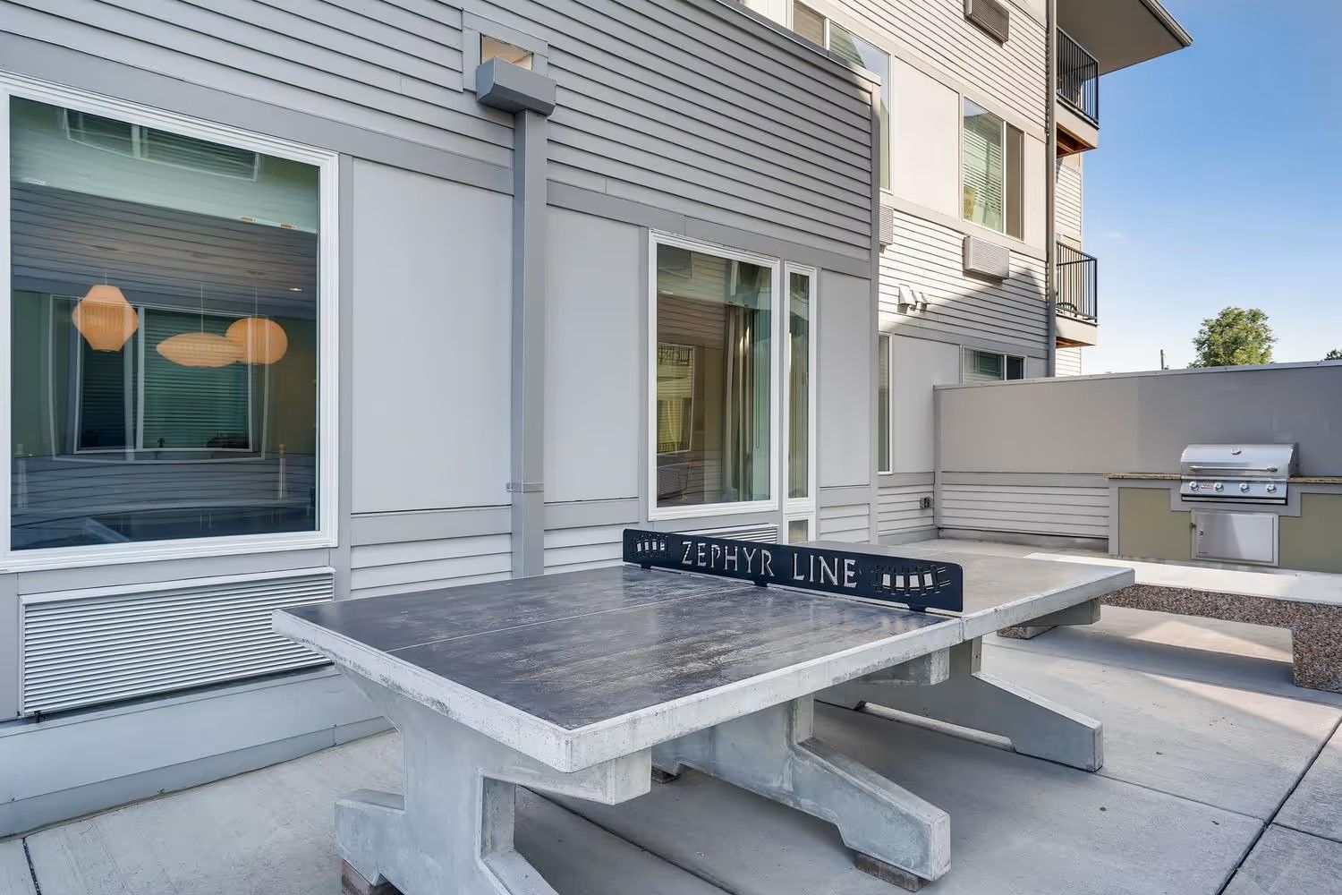 A concrete outdoor ping-pong table on a patio next to a modern building with siding and a built-in grill.
