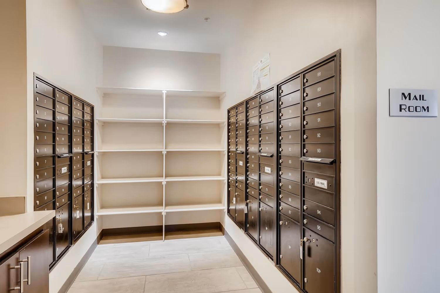 A mail room featuring two rows of metal mailboxes and center shelving on a light-colored tile floor.