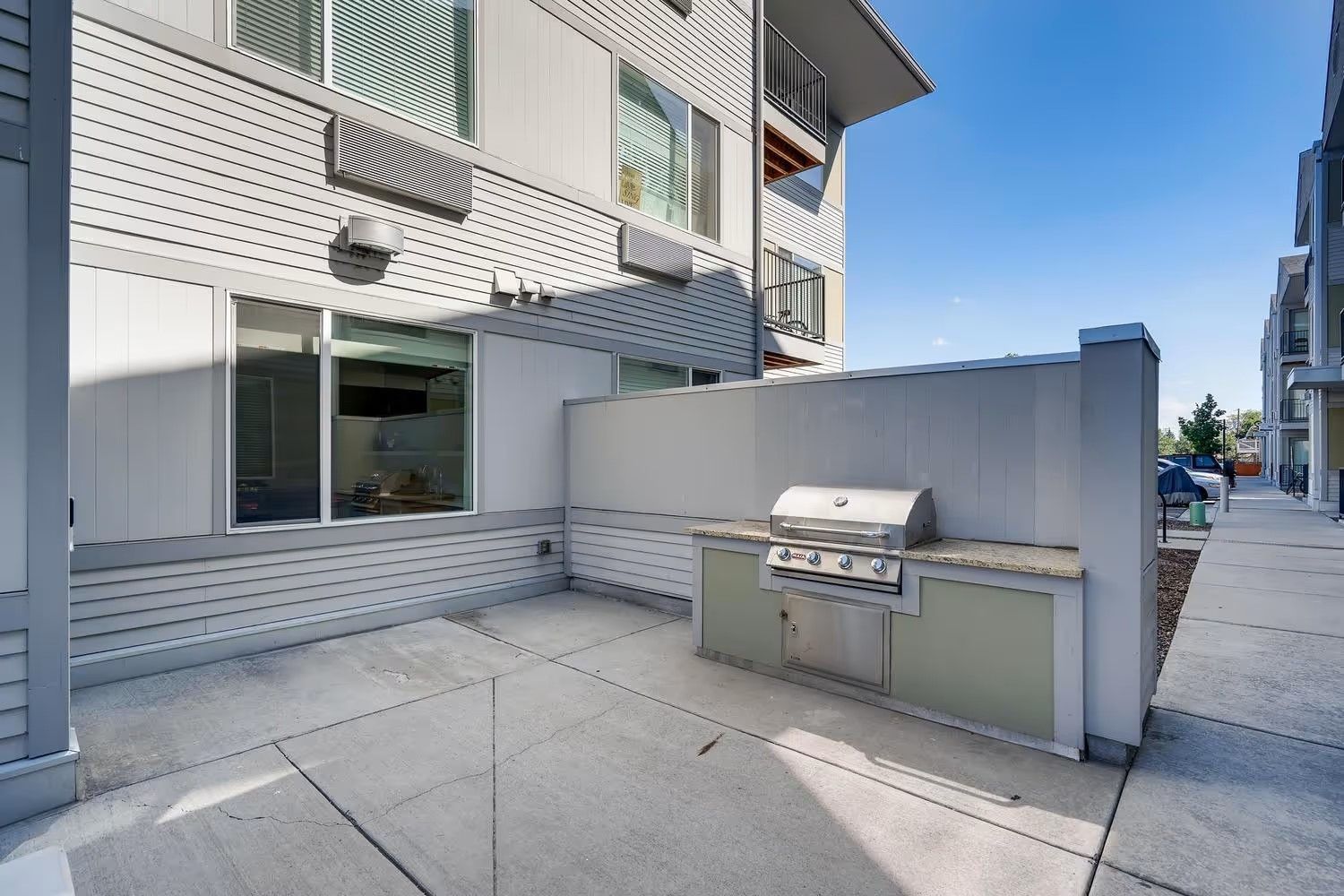 Outdoor patio with a built-in stainless steel grill set against a light grey building wall under a clear blue sky.
