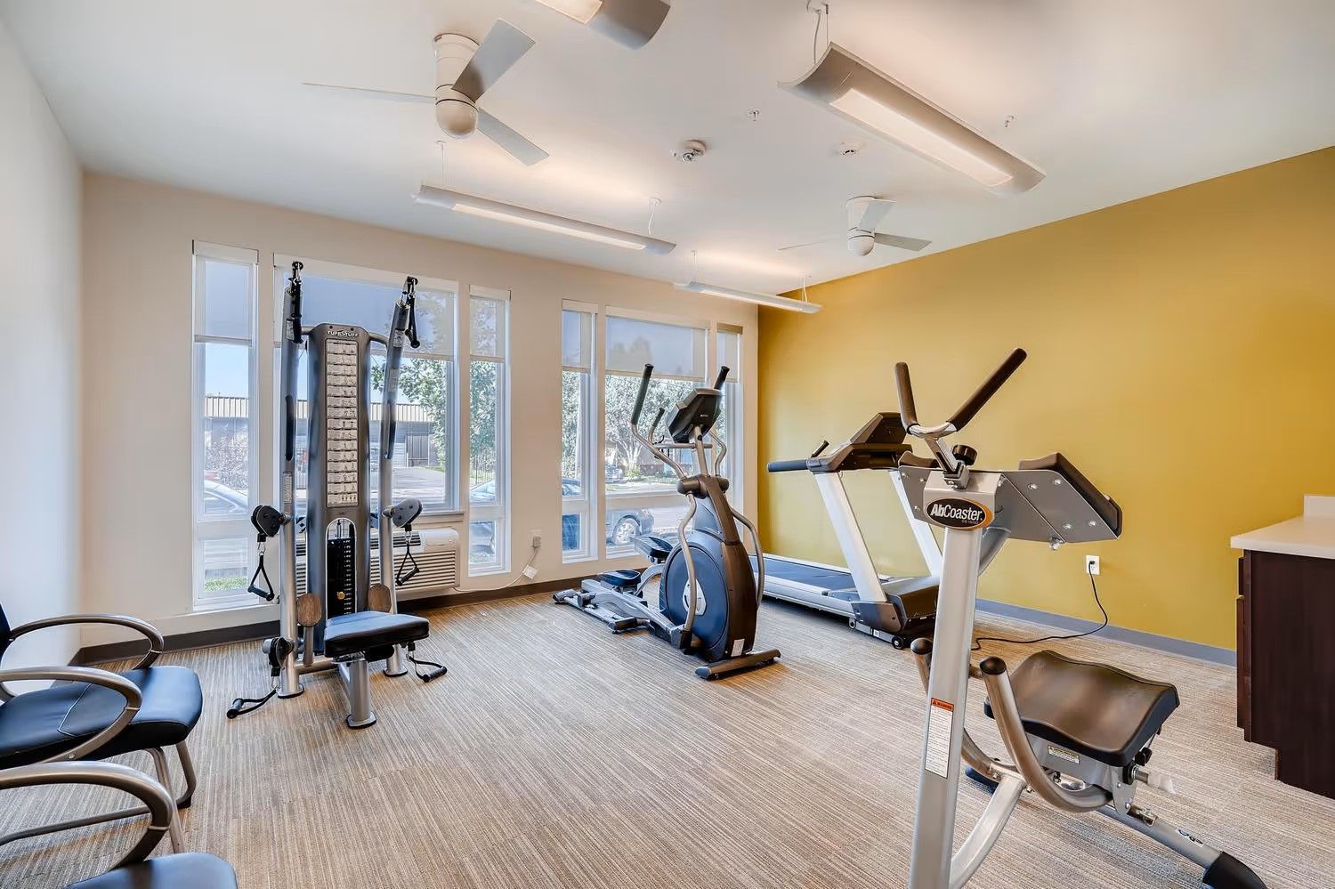 A bright gym with a multi-station weight machine, an elliptical, a treadmill, and a chair against a yellow wall.