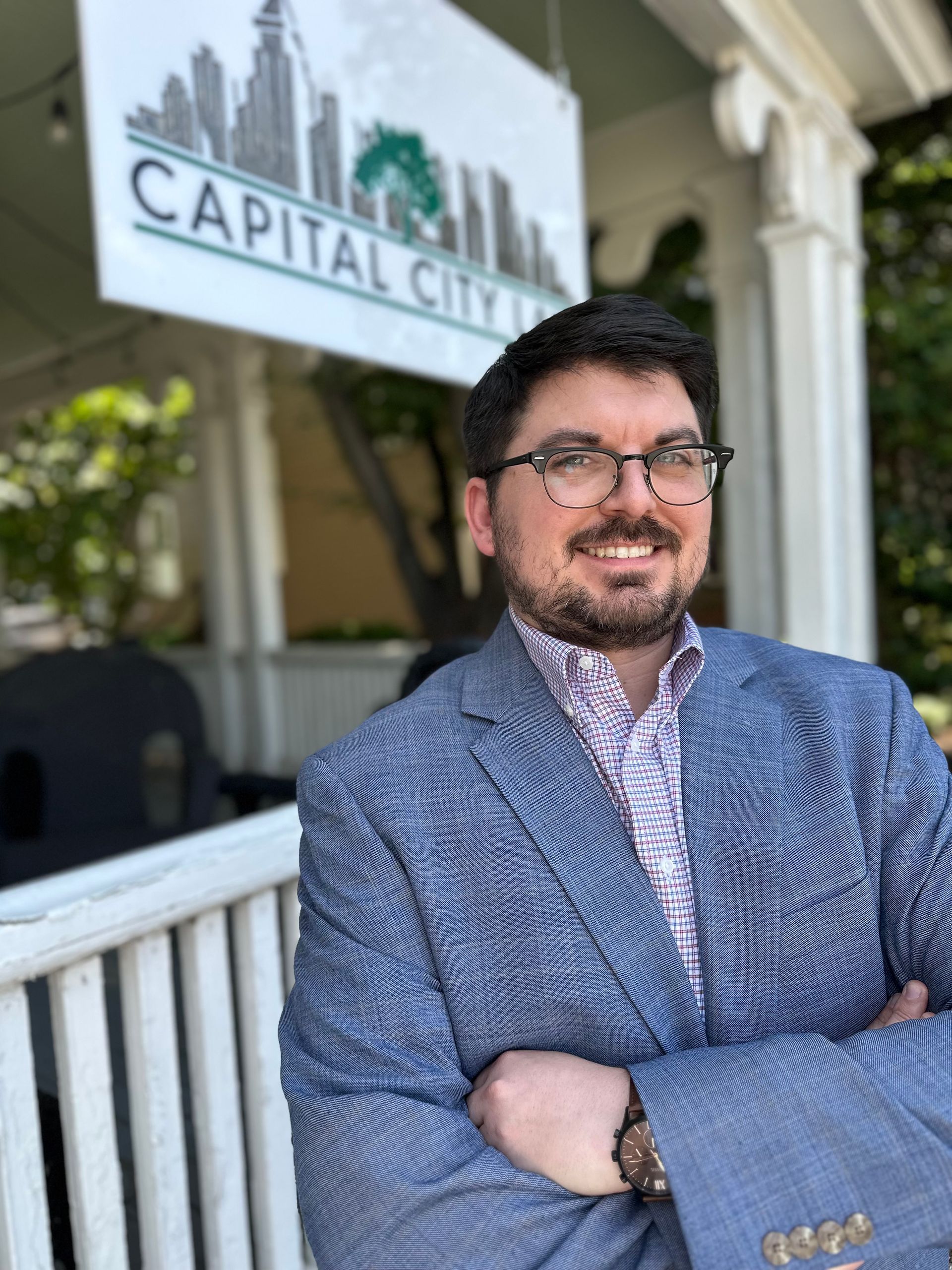 Man in glasses, blue blazer smiles outside Capital City Inn sign.