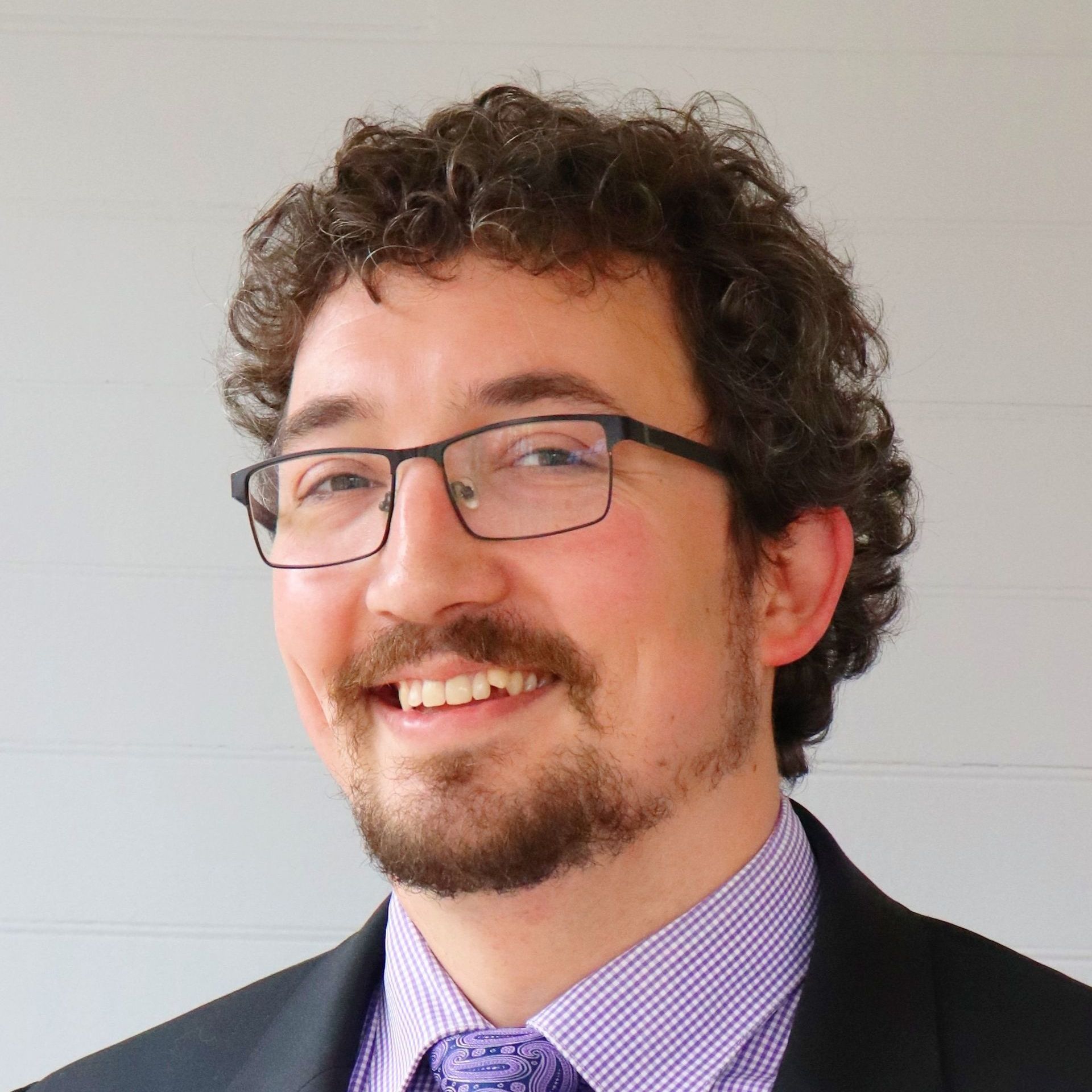 Man with curly brown hair, glasses, and beard smiles, wearing a purple patterned tie, and a black suit.