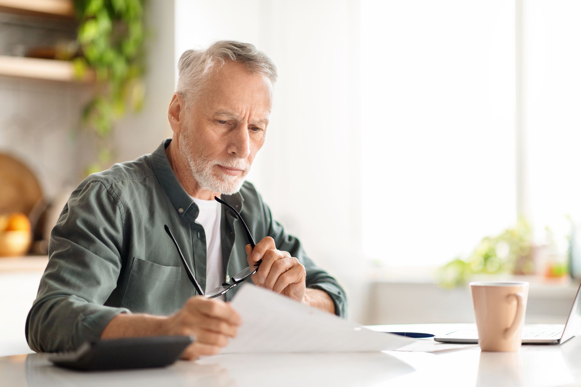 Man looking at documents, holding glasses, sitting at a desk with a calculator, coffee, and laptop.