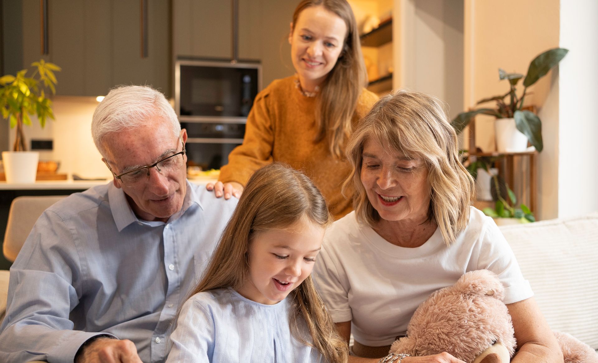 Family reads together on a couch, a girl smiles at a book. Others watch: grandparents and an older girl.
