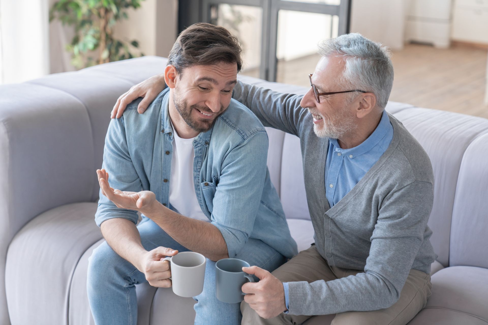 Two men on a sofa, one with arm around the other's shoulder, smiling and holding mugs.