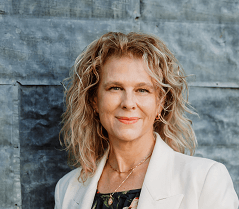 Portrait of a smiling person in a white blazer against a textured blue-gray wall