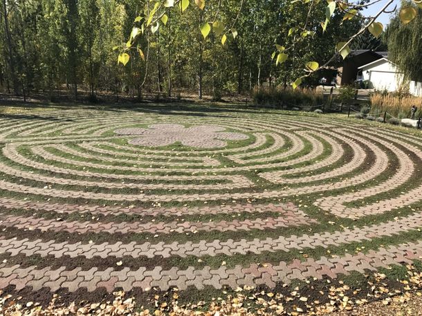 Circular grassy labyrinth in a park, bordered by trees and a house in the background.