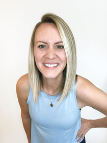 Smiling blonde person in a light blue sleeveless top against a white background, hand on hip