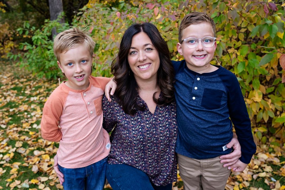 Three people smiling outdoors in autumn, with two children standing beside a woman.