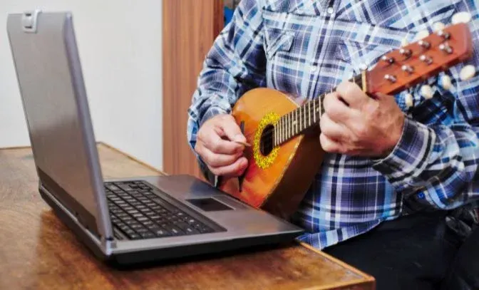 Person playing a small orange ukulele beside an open laptop on a wooden table
