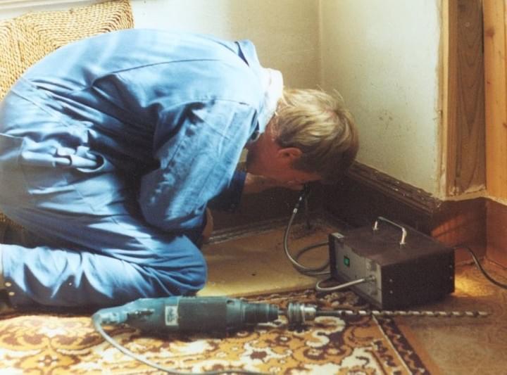 Eine Person in blauem Overall kniet und untersucht mit einem Gerät und einer Bohrmaschine auf dem Boden eine Ecke.