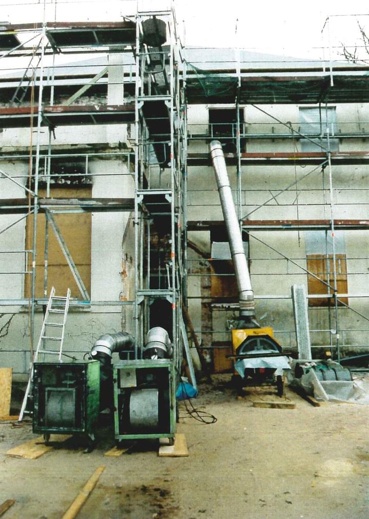 Baumaschinen auf einem Gerüst neben einem Gebäude. Ein hohes Metallförderband ist mit einer Maschine verbunden.
