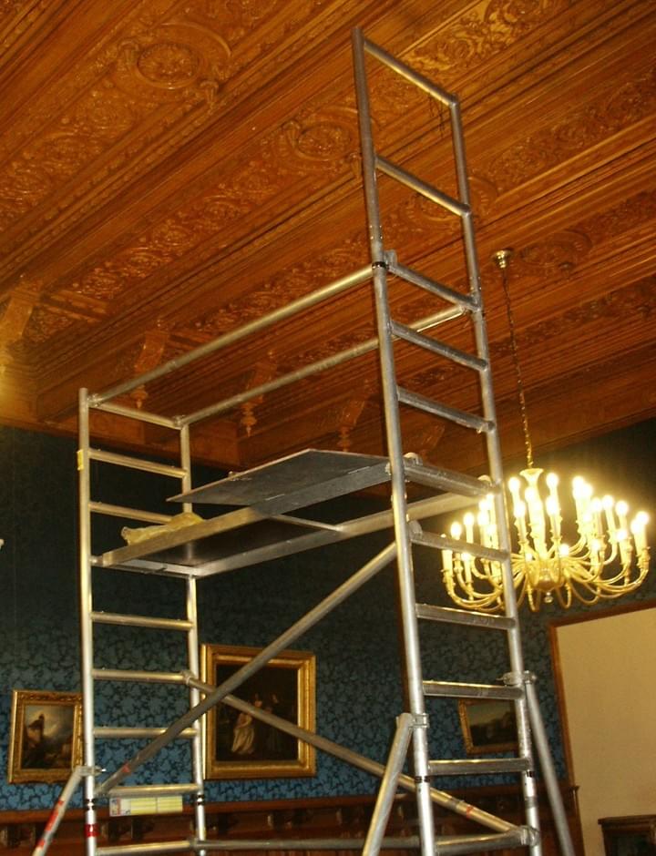 Ein Gerüst in einem Raum mit dekorativer Holzdecke, kunstvollem Kronleuchter und Kunstwerken an den Wänden.