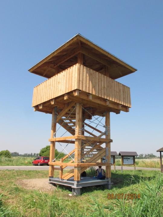 Hölzerner Aussichtsturm auf einer Wiese; Person sitzt unten, rotes Auto auf der Straße zu sehen, blauer Himmel.