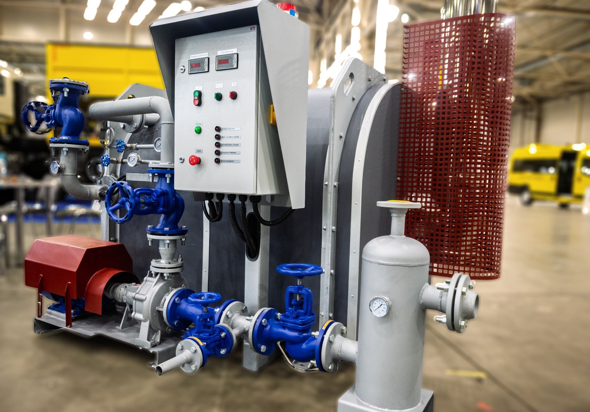 Industrial pump and pipe system with control panel, valves, and red mesh guard in a factory setting