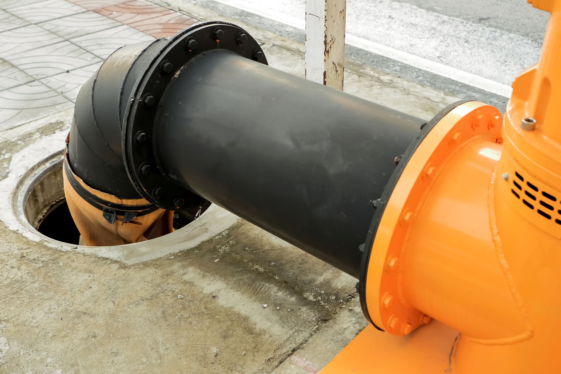 Black pipe connected to orange industrial equipment beside a concrete drain opening