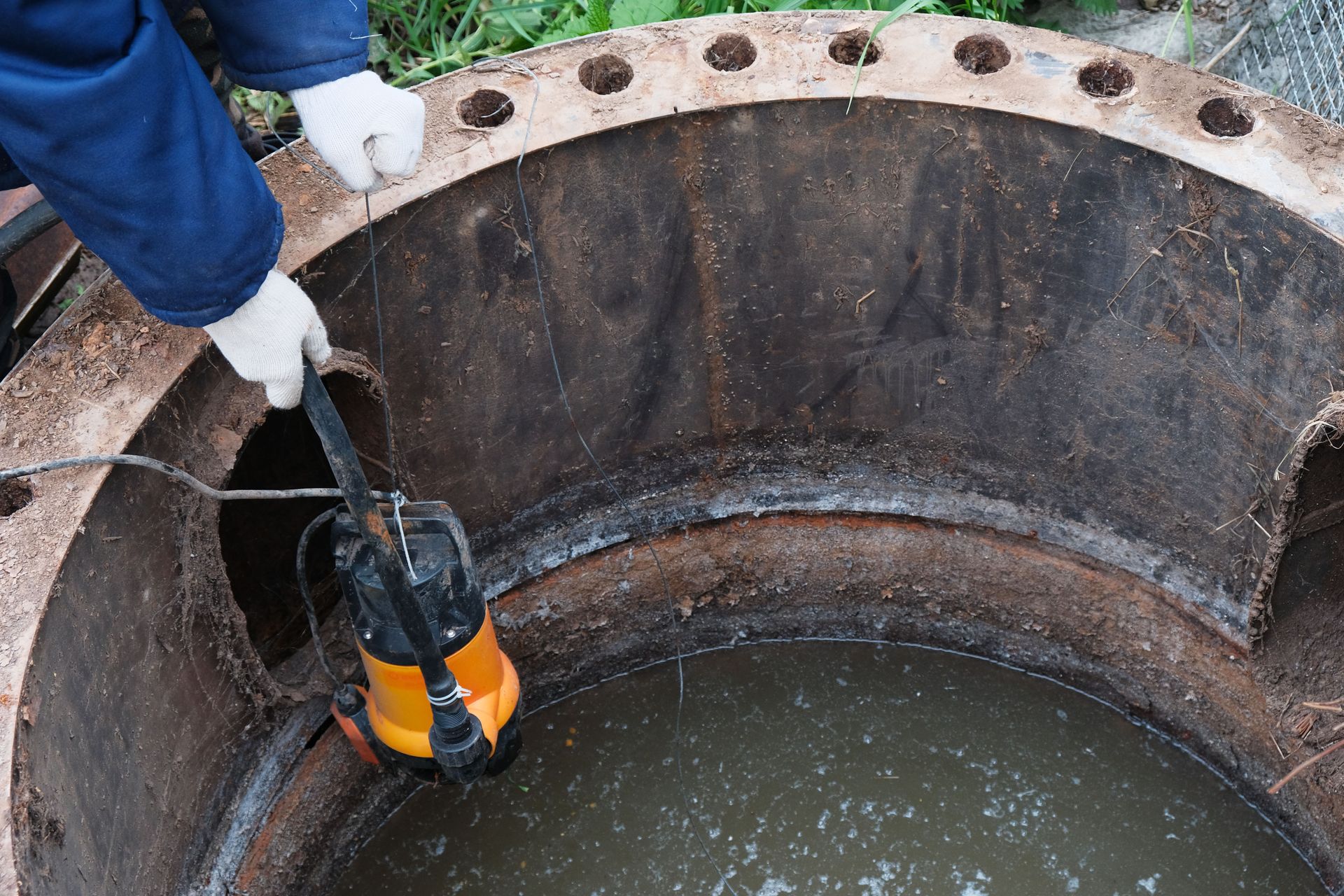 Worker lowering a yellow pump into a deep concrete manhole filled with water