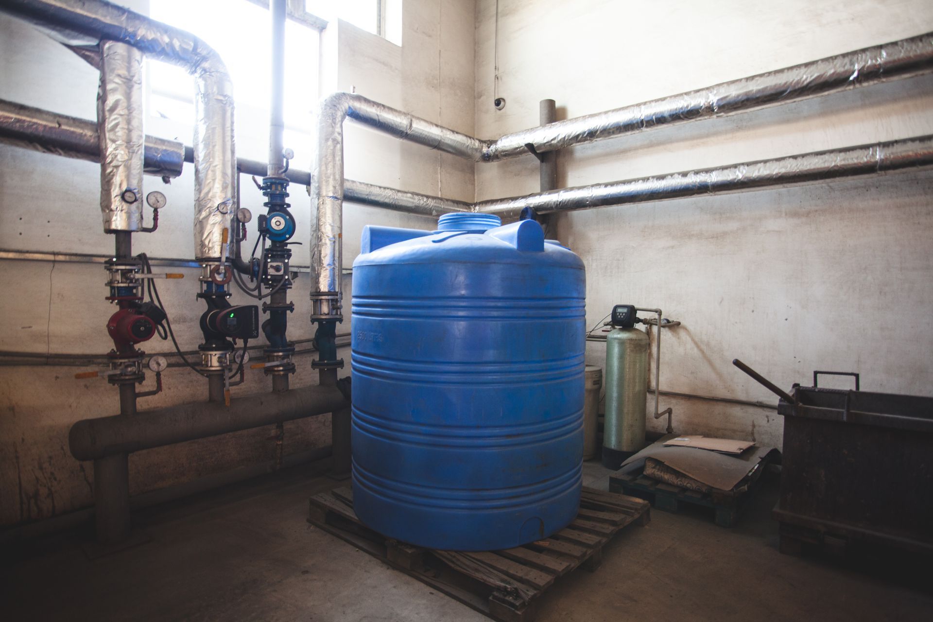 Utility room with a large blue water tank, pipes, valves, and equipment against concrete walls.