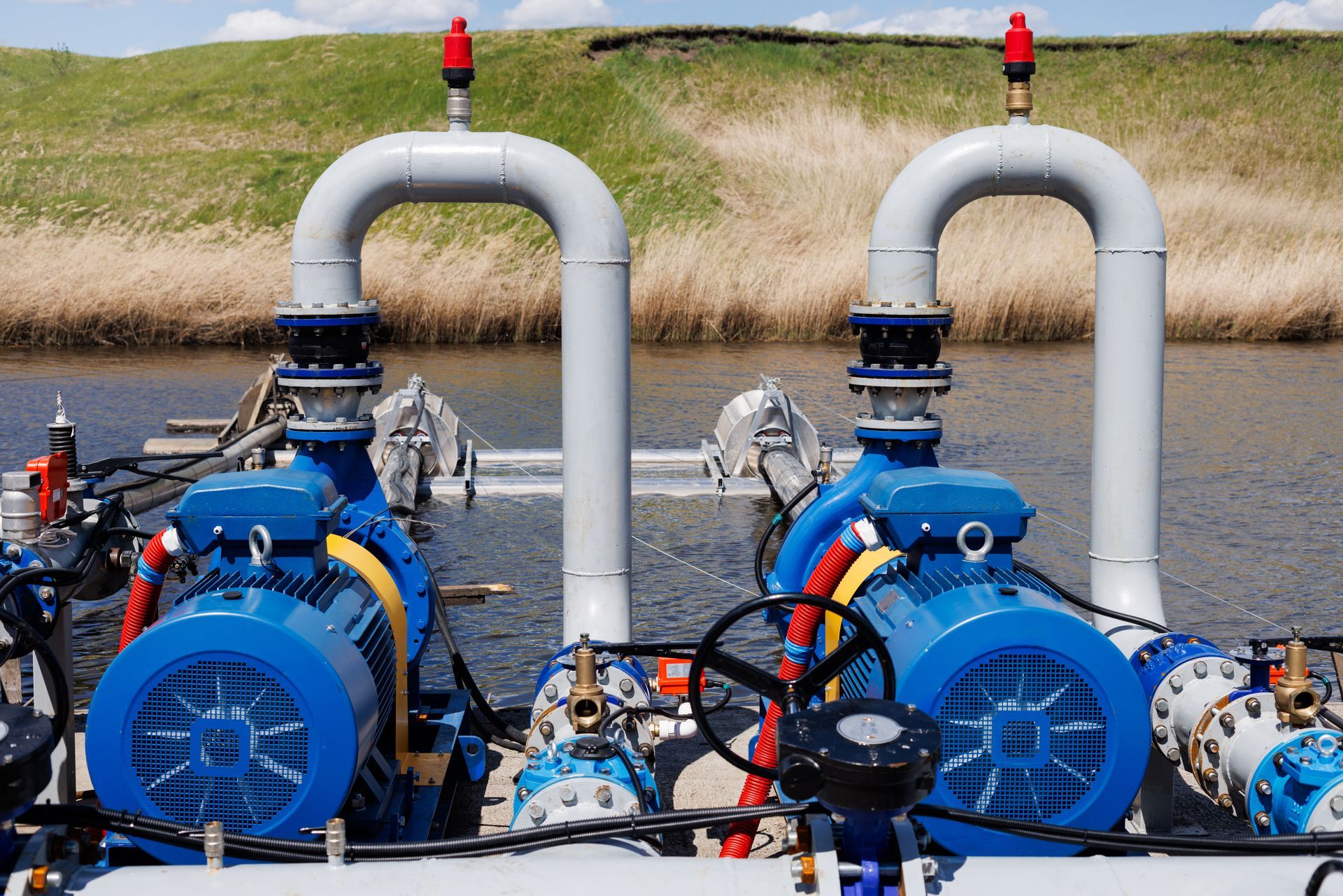 Blue industrial pumps and pipes beside a body of water, with grassy banks in the background