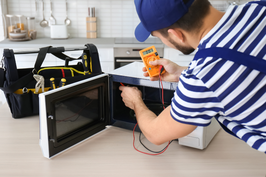 A man is fixing a microwave with a multimeter in a kitchen.