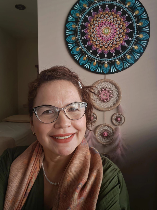 Una persona sonriente con gafas y bufanda en una habitación, con un colorido mural de mandalas detrás.