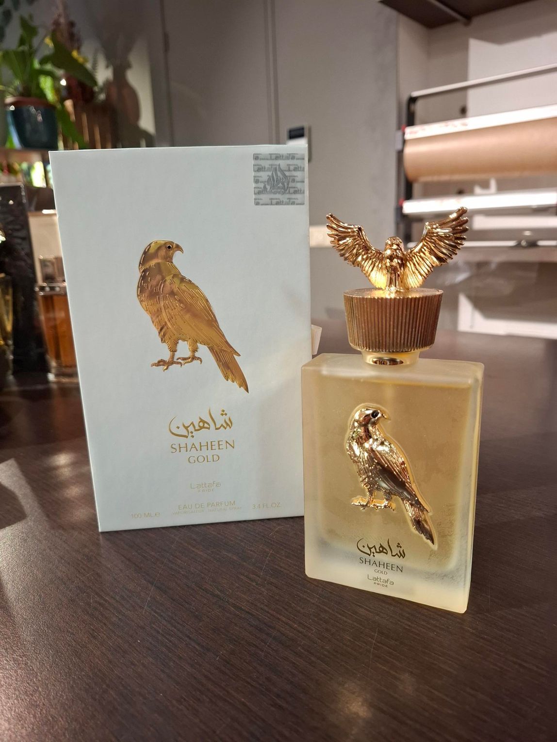 Een flesje Shaheen Gold-parfum met een gouden valkembleem en bijpassende dop staat naast de witte doos.