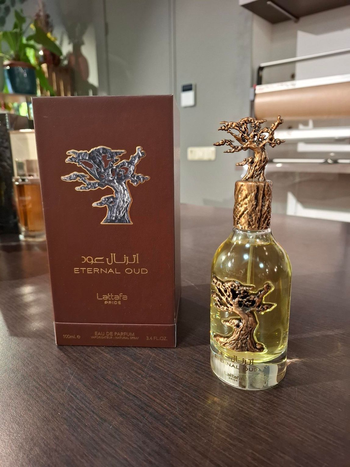 De flacon van het parfum Eternal Oud en de bijbehorende bruine verpakking, met een gouden boommotief, staan op een donkere houten tafel.
