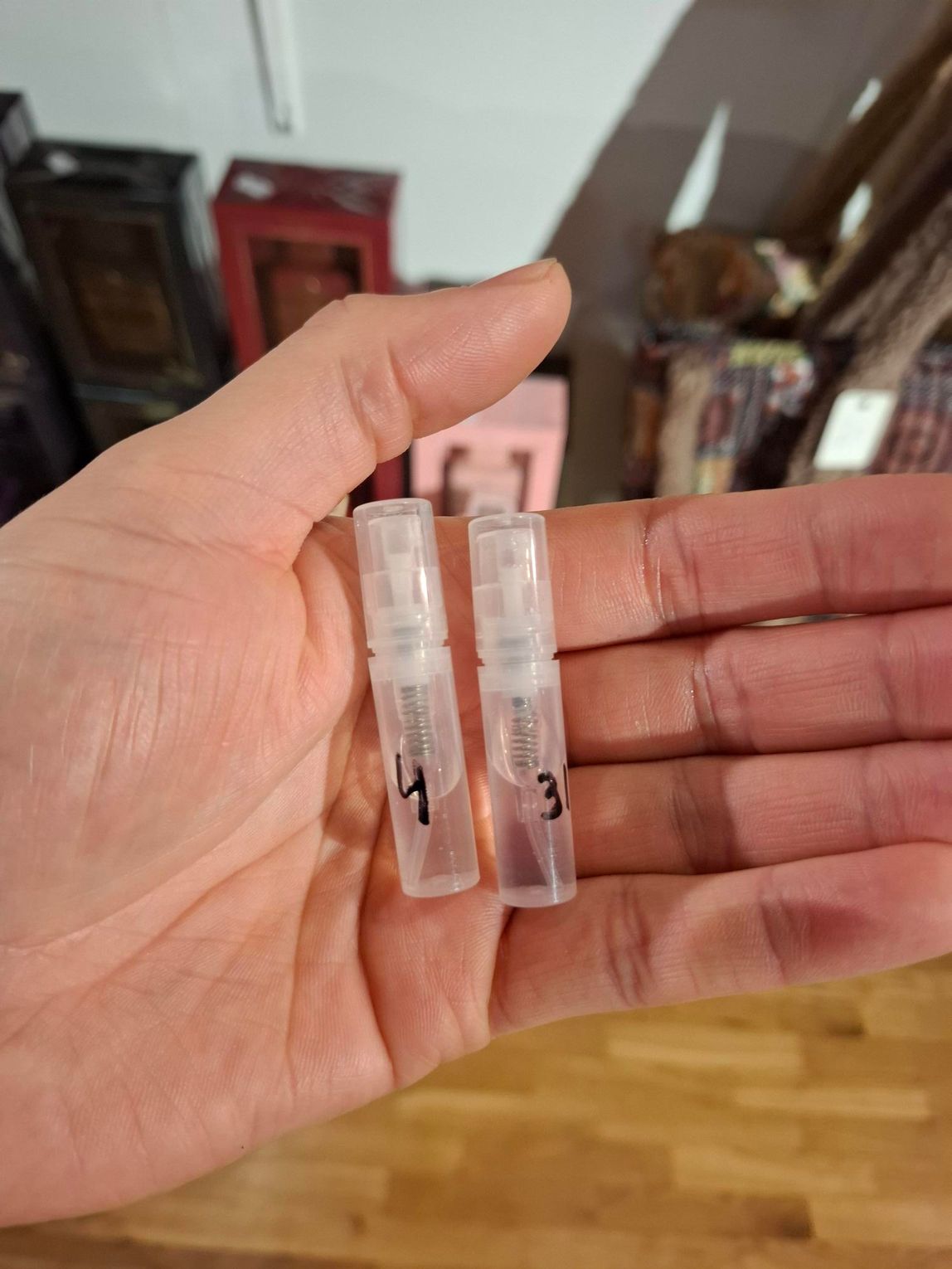 Een hand die twee kleine, doorzichtige plastic flesjes met zwarte markeringen vasthoudt, binnen op een houten vloer.