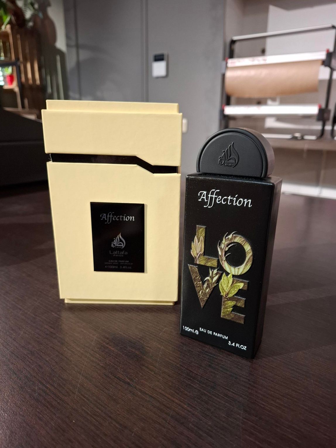 Een flesje parfum Affection Love en het lichtgekleurde doosje staan op een donkerhouten ondergrond.