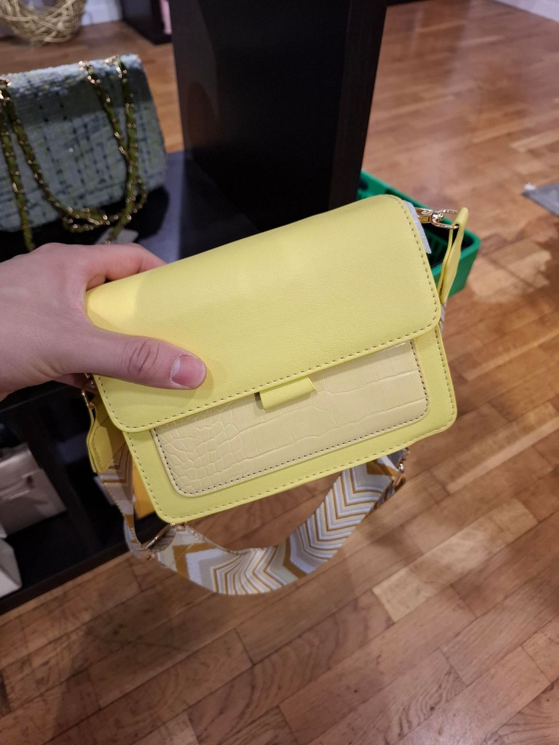 Hand die een klein geel handtasje vasthoudt boven een houten vloer in een winkel.