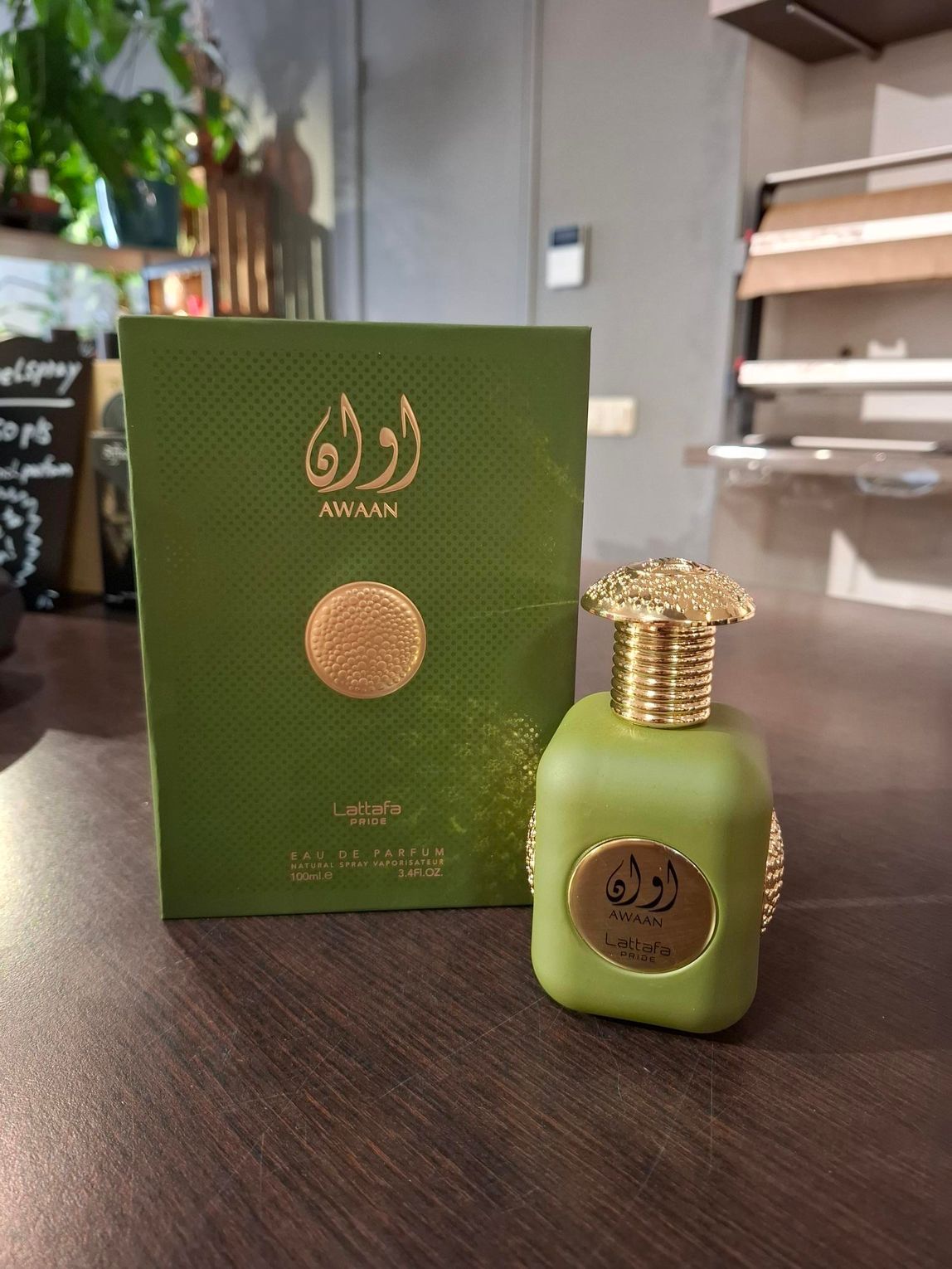 Een saliegroene parfumfles en de bijbehorende rechthoekige doos met gouden Arabische kalligrafie, geplaatst op een houten ondergrond.