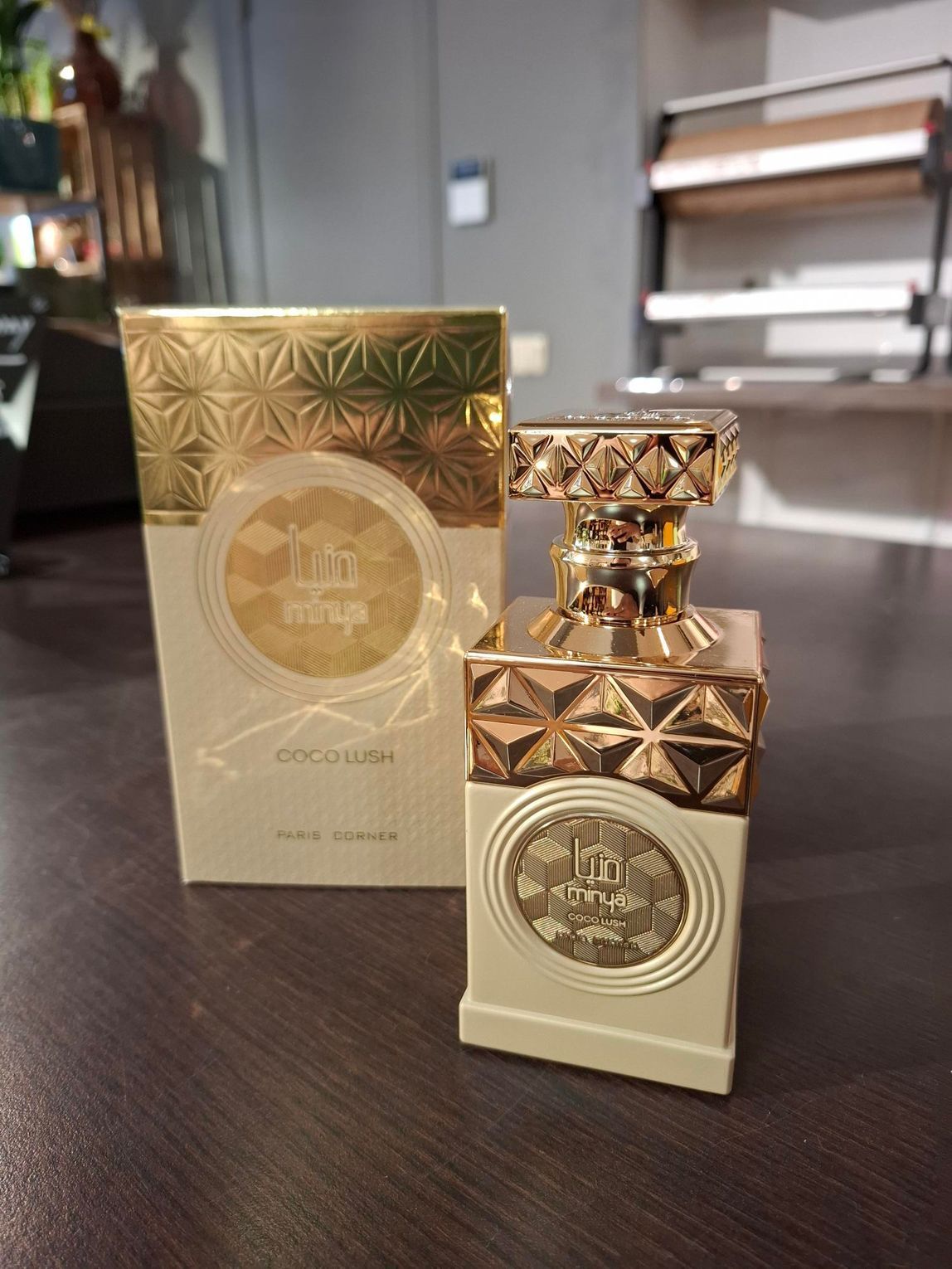 Een flesje beige en goudkleurige parfum met een bijpassende doos met patroon, staand op een donkerhouten ondergrond.