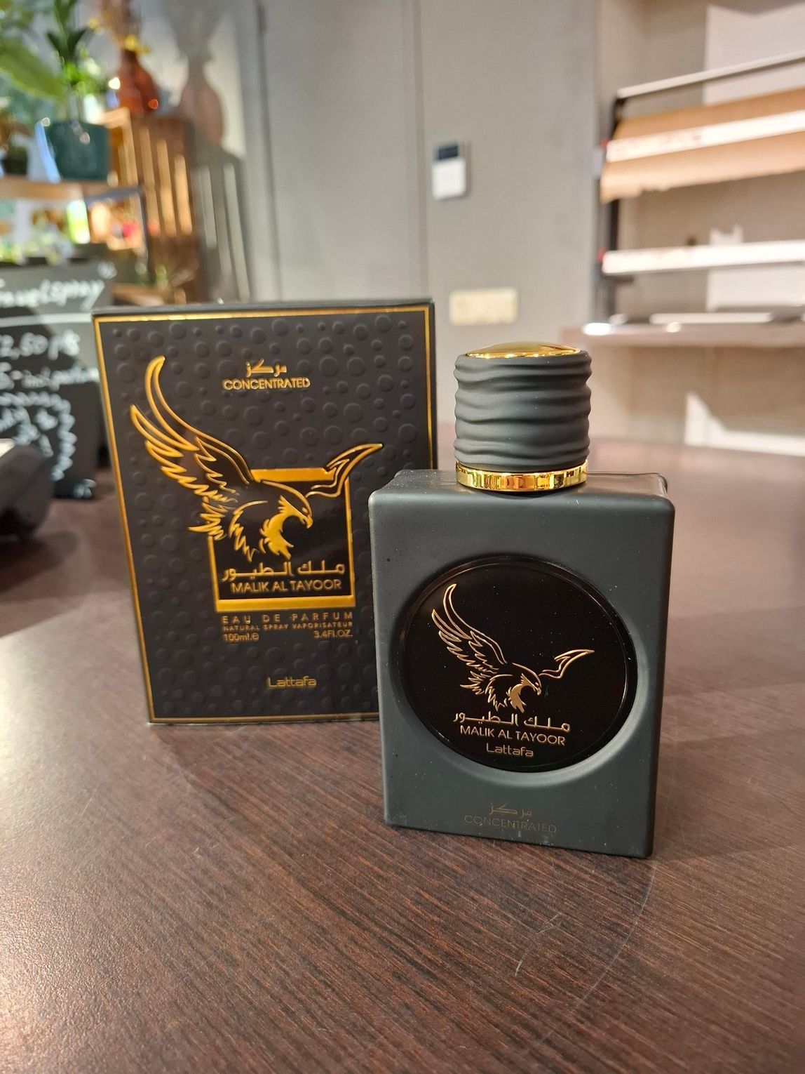 Een donkergrijze fles Lattafa Ameer Al Oudh parfum met een gouden adelaarslogo, naast de bijpassende, gestructureerde presentatiedoos.