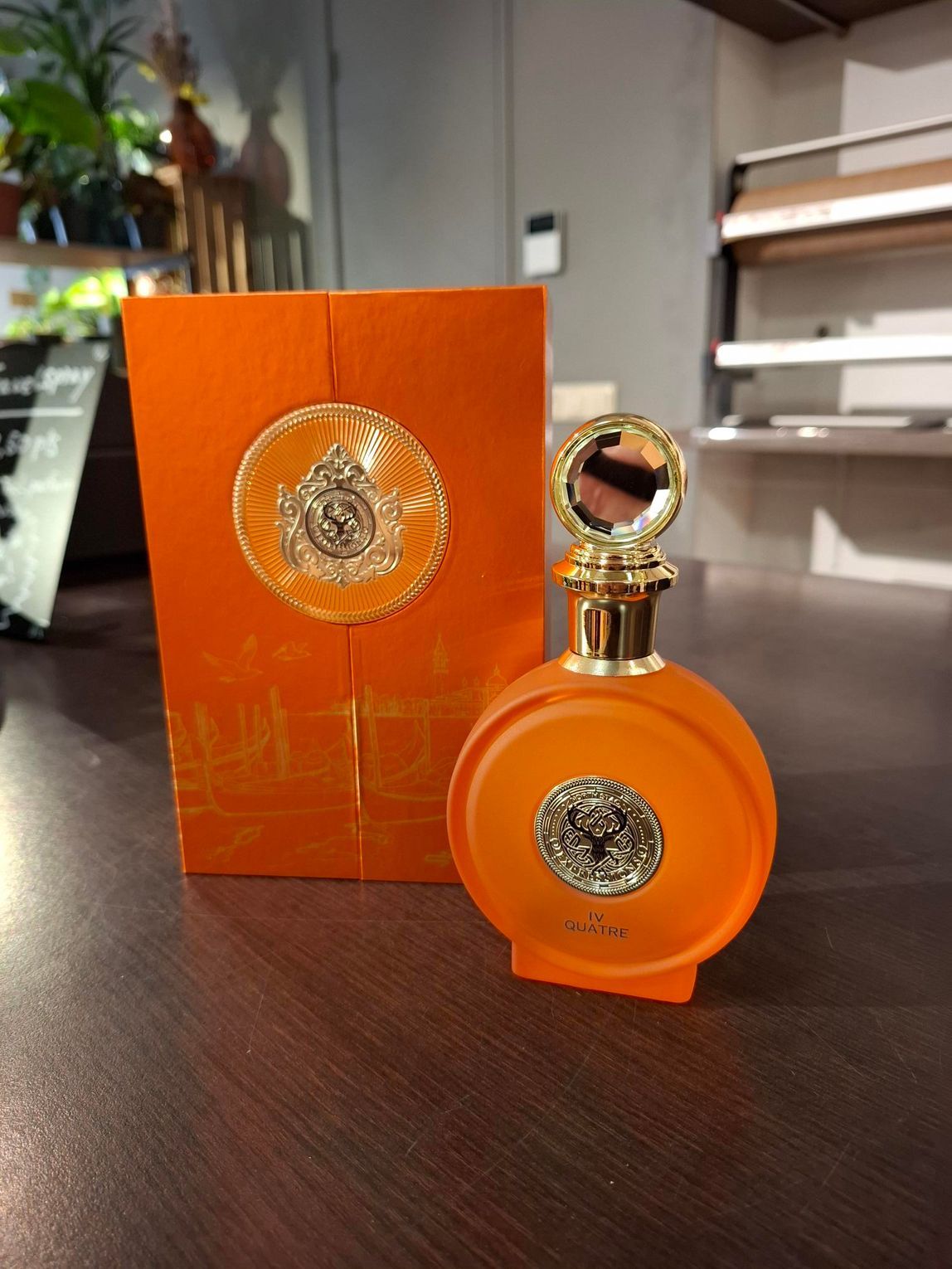 Een oranje, ronde parfumfles met een gouden embleem staat naast de bijpassende oranje doos op een donkerhouten ondergrond.