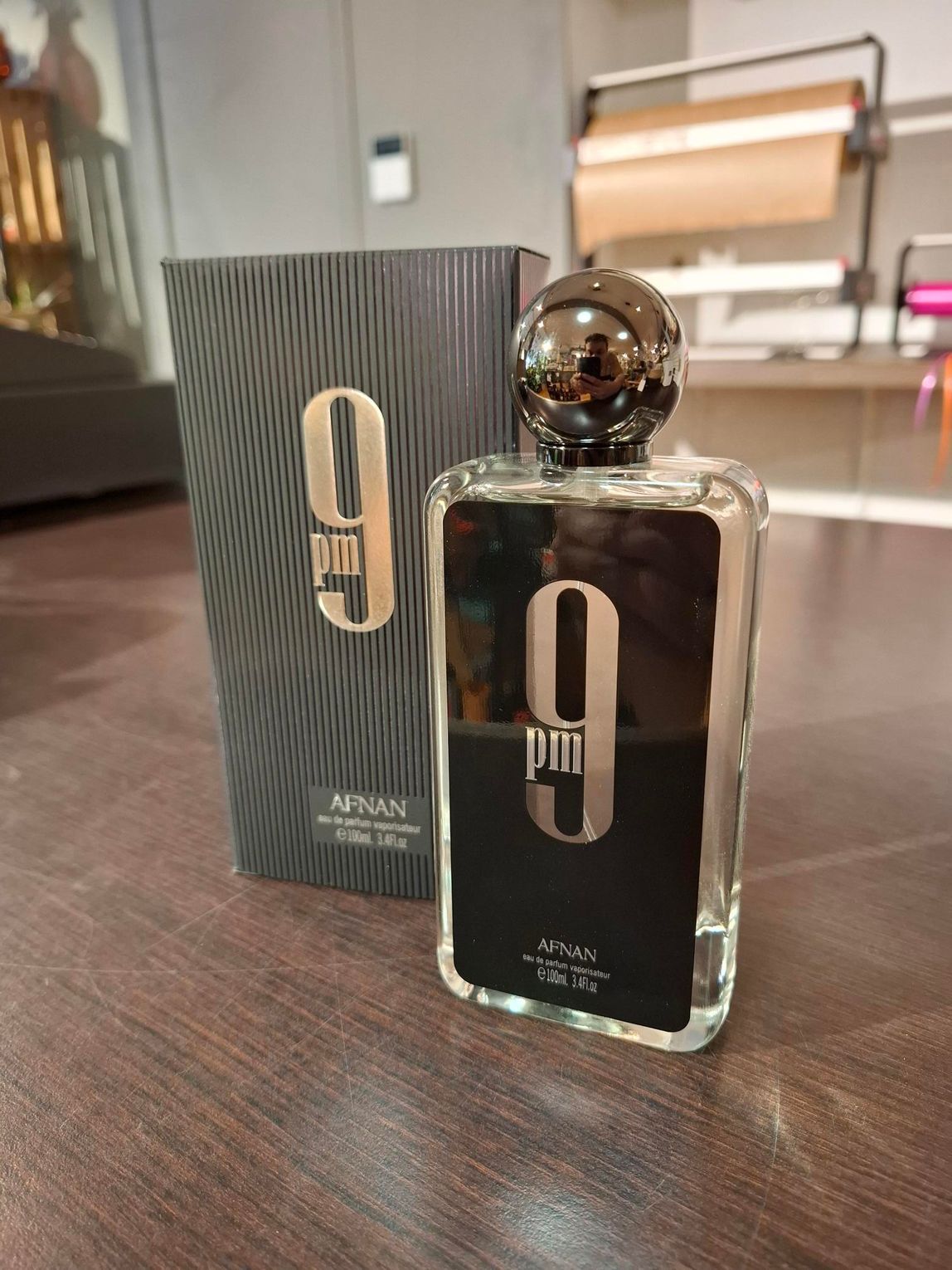 Een fles Afnan 9 PM eau de cologne en de bijbehorende zwarte doos met patroon staan op een houten ondergrond.
