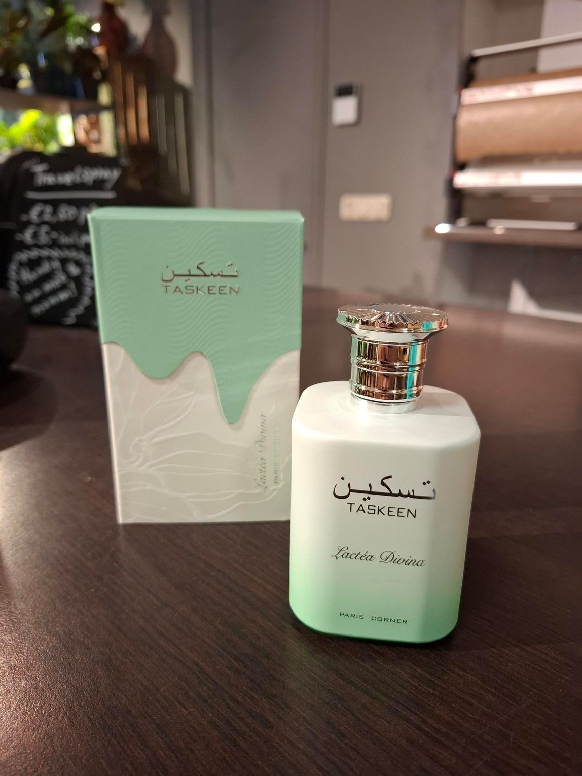 Een flesje Taskeen-parfum met de bijbehorende lichtgroene en witte doos, staand op een houten ondergrond.