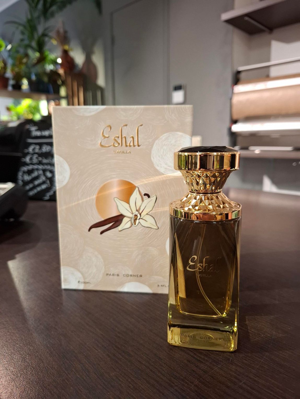 Een flesje Eshal-parfum naast de beige doos met een afbeelding van een vanillebloem op een donkere houten tafel.