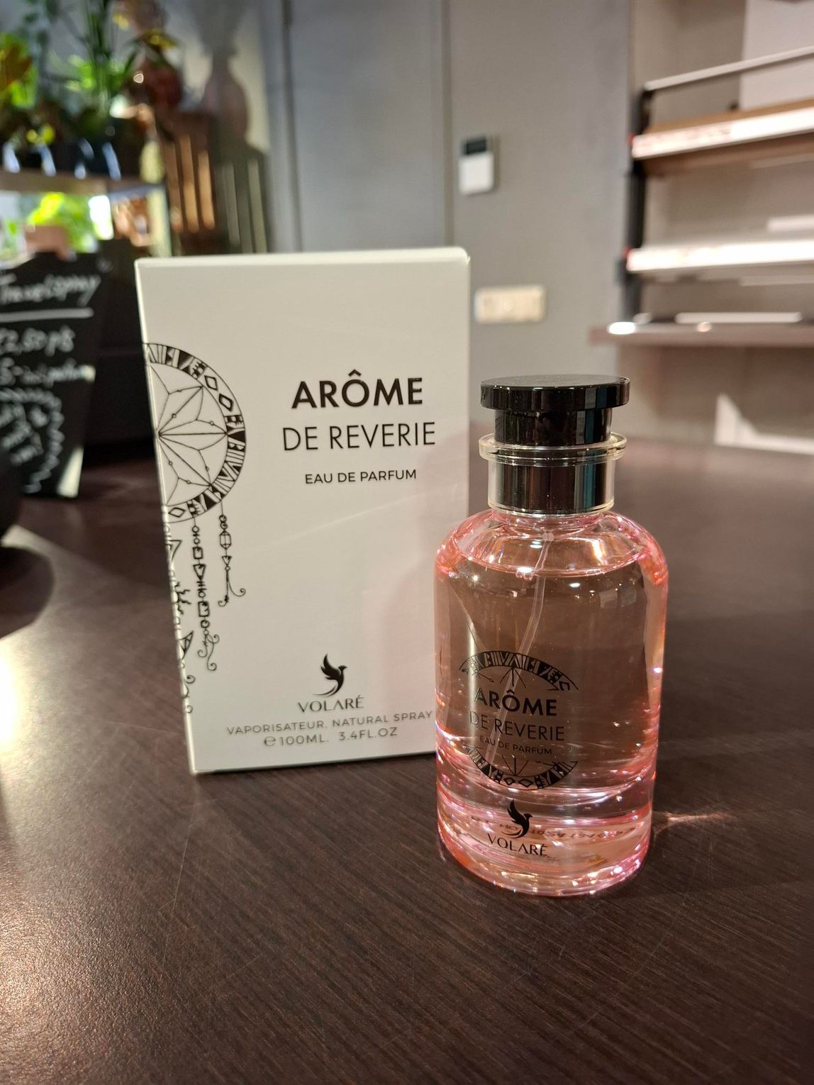 Een flesje roze parfum Arôme de Reverie en de bijbehorende witte verpakking staan op een donkerhouten oppervlak in een winkel.