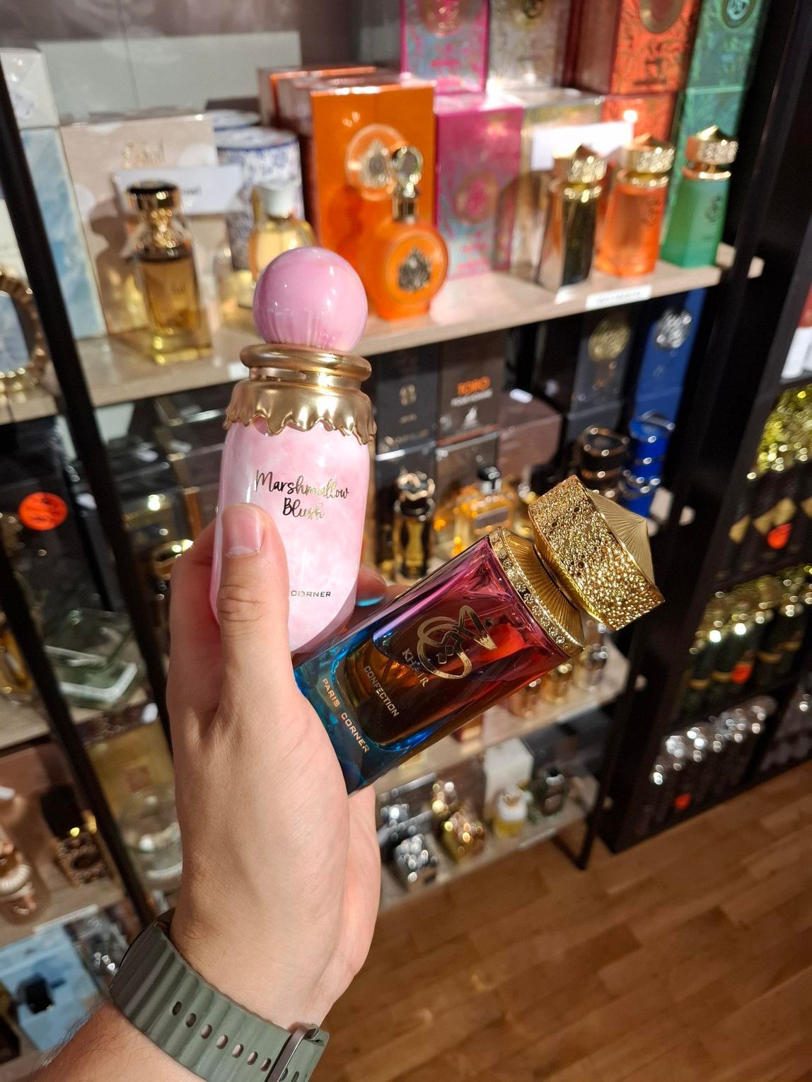 Een hand houdt twee roze parfumflesjes vast voor een plank vol met verschillende geuren.