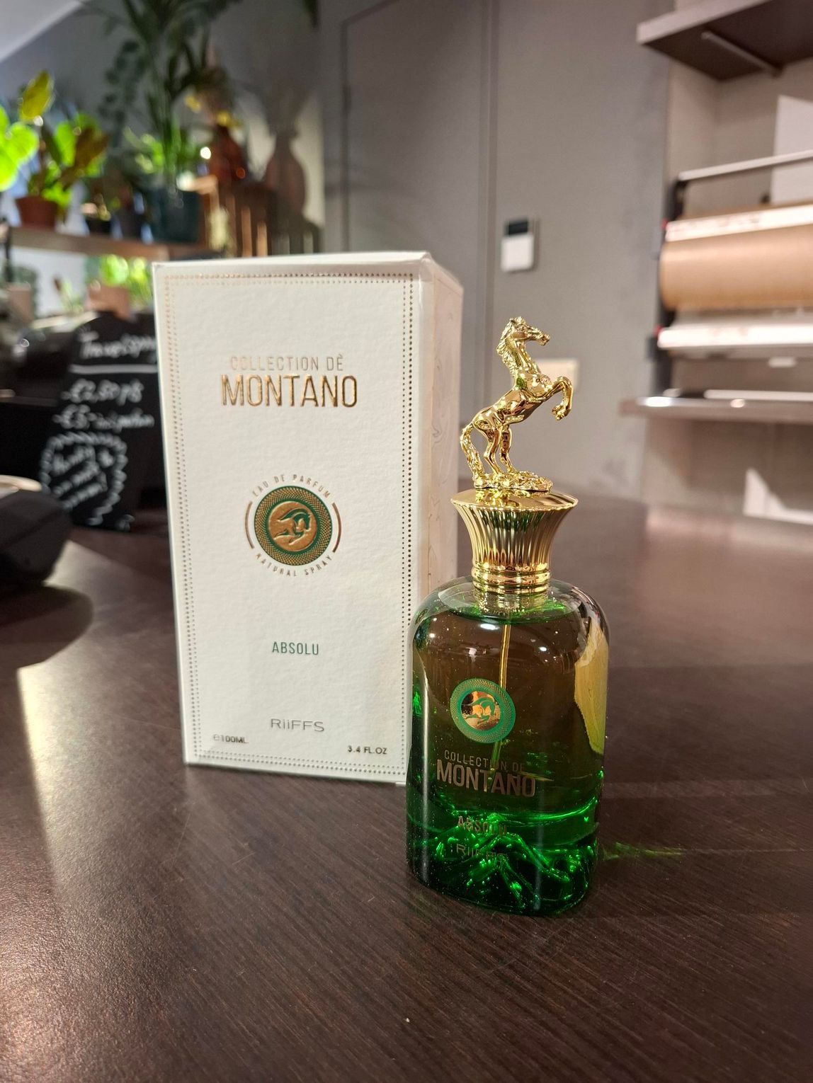 Een flesje Montano-parfum met een gouden paardendop naast de witte doos, die op een donkerhouten ondergrond staat.