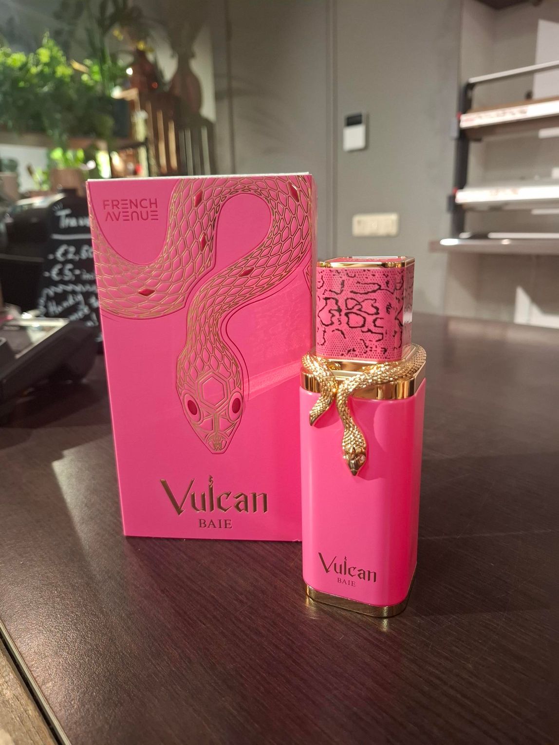 Een felroze parfumflesje en het bijbehorende doosje, beide versierd met gouden slangenmotieven, staan op een houten toonbank.