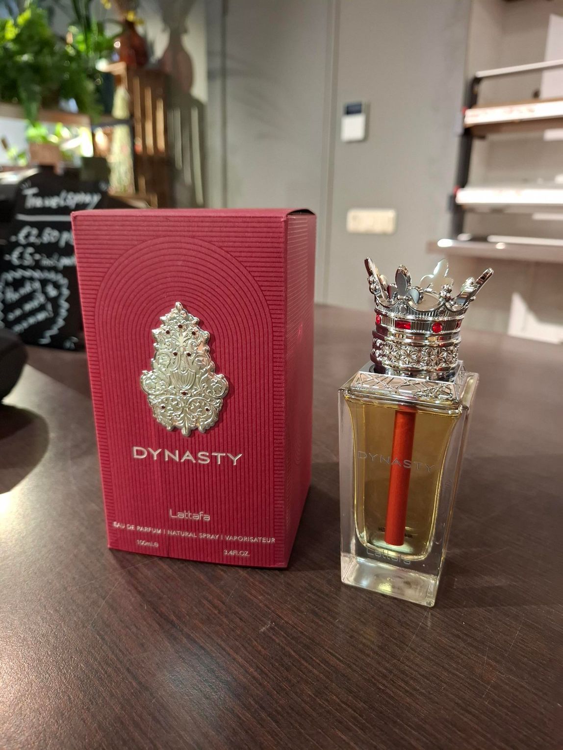 Een parfumflesje van Dynasty en de bijbehorende rode, gestructureerde doos met een sierlijk zilveren embleem, staan op een houten ondergrond.