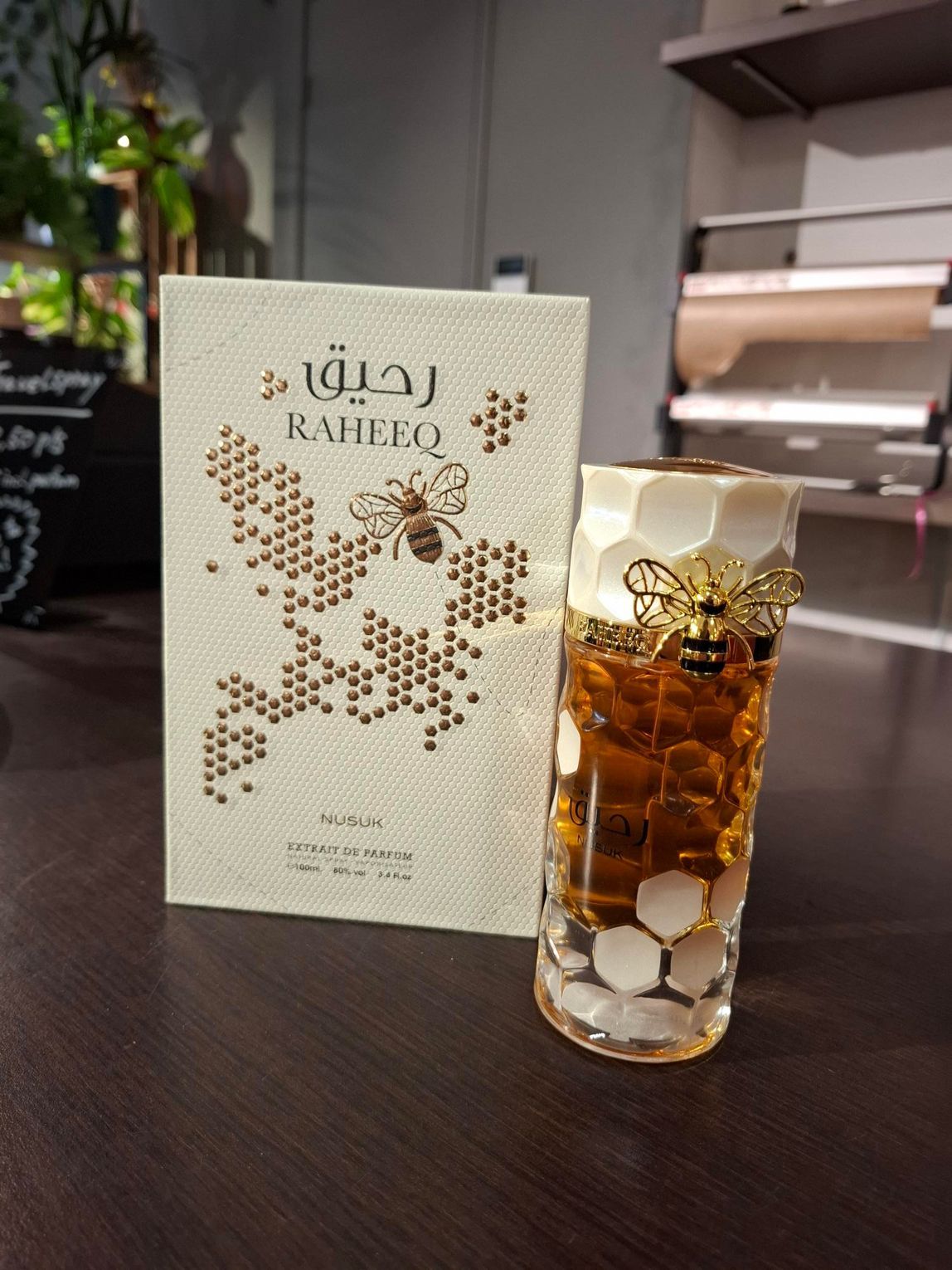 Een fles Raheeq-parfum en de bijbehorende doos, beide met honingraatpatroon en een gouden bijenornament, op een donkere tafel.