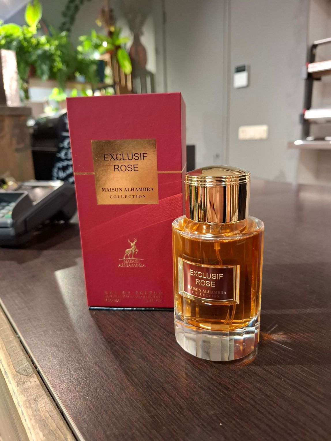 Een fles Exclusive Rose-parfum en de bijbehorende rode doos staan op een donkerhouten ondergrond in een binnenruimte.