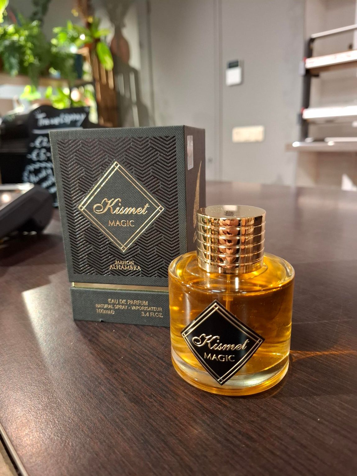 Een flesje Kismet Magic-parfum en de bijbehorende zwarte, gestructureerde doos staan op een donkere houten tafel in een kamer met planten.