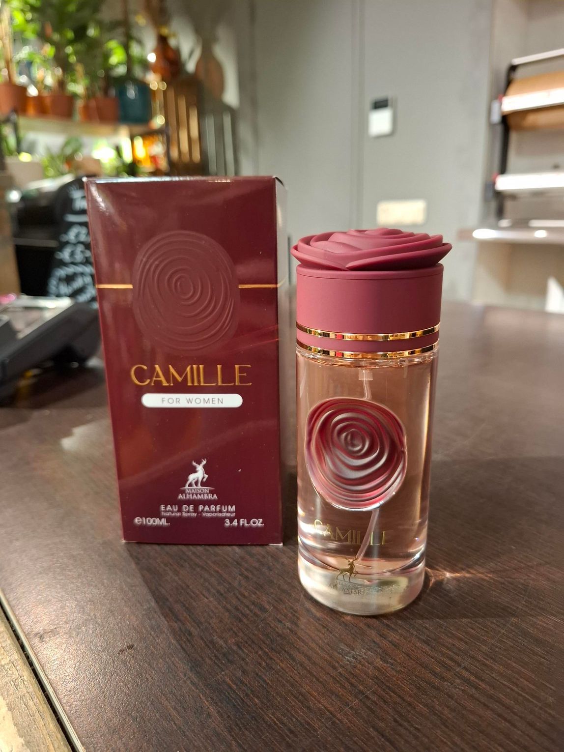 Een flesje Camille Eau de Parfum en de bijbehorende bordeauxrode doos met rozenembleem, staan op een houten ondergrond.