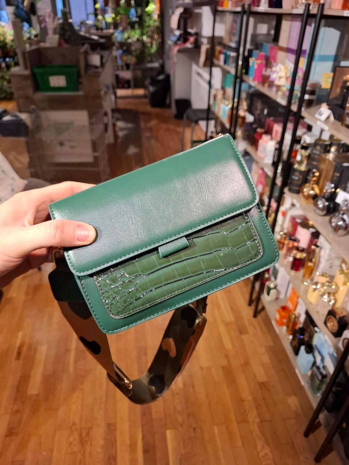 Hand die een kleine groene leren portemonnee vasthoudt in een winkelgang met schappen vol koopwaar.